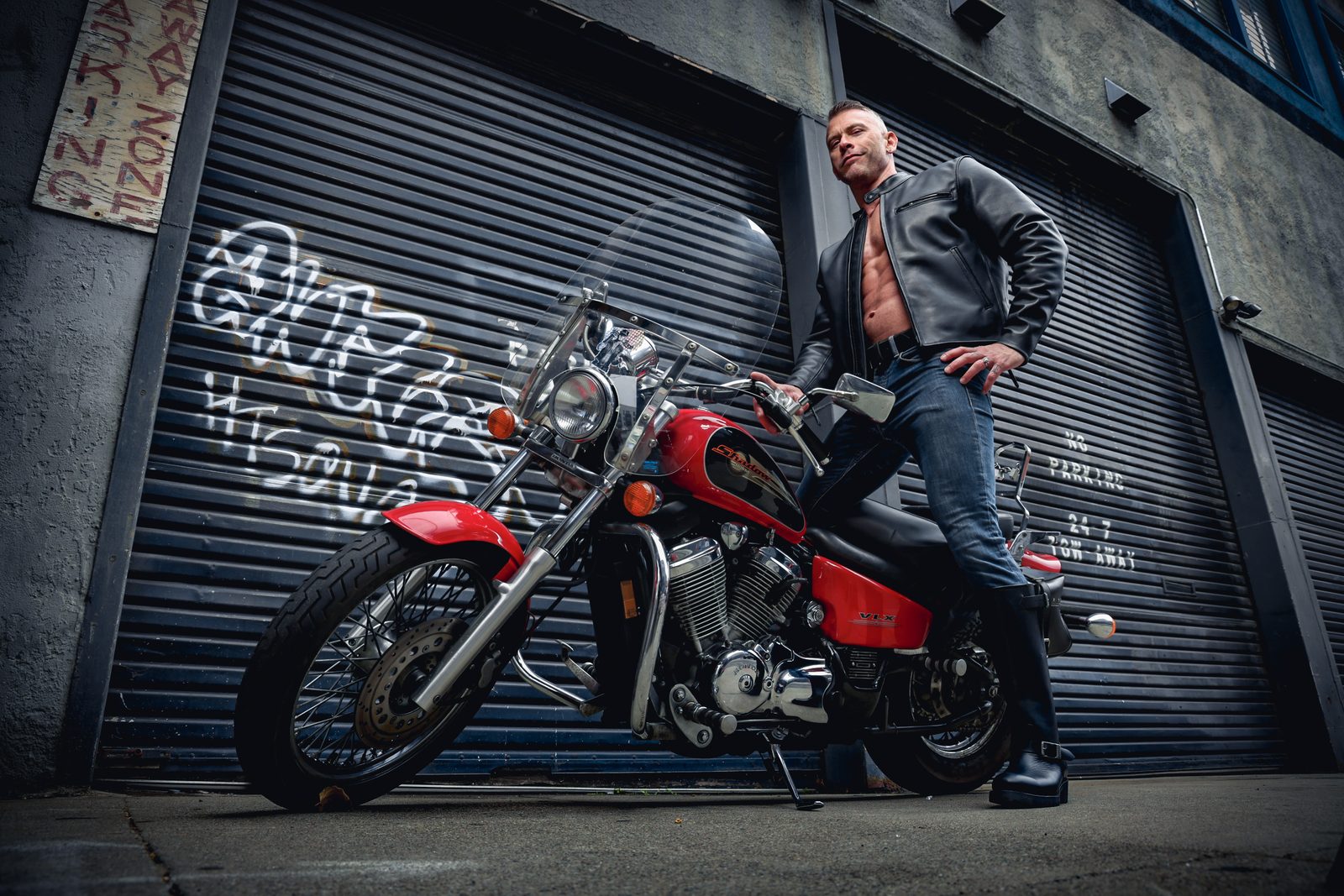 On location: a man in a leather jacket astride a red motorcycle in an SF alley of graffitied roll-up doors, shot from a low heroic angle.