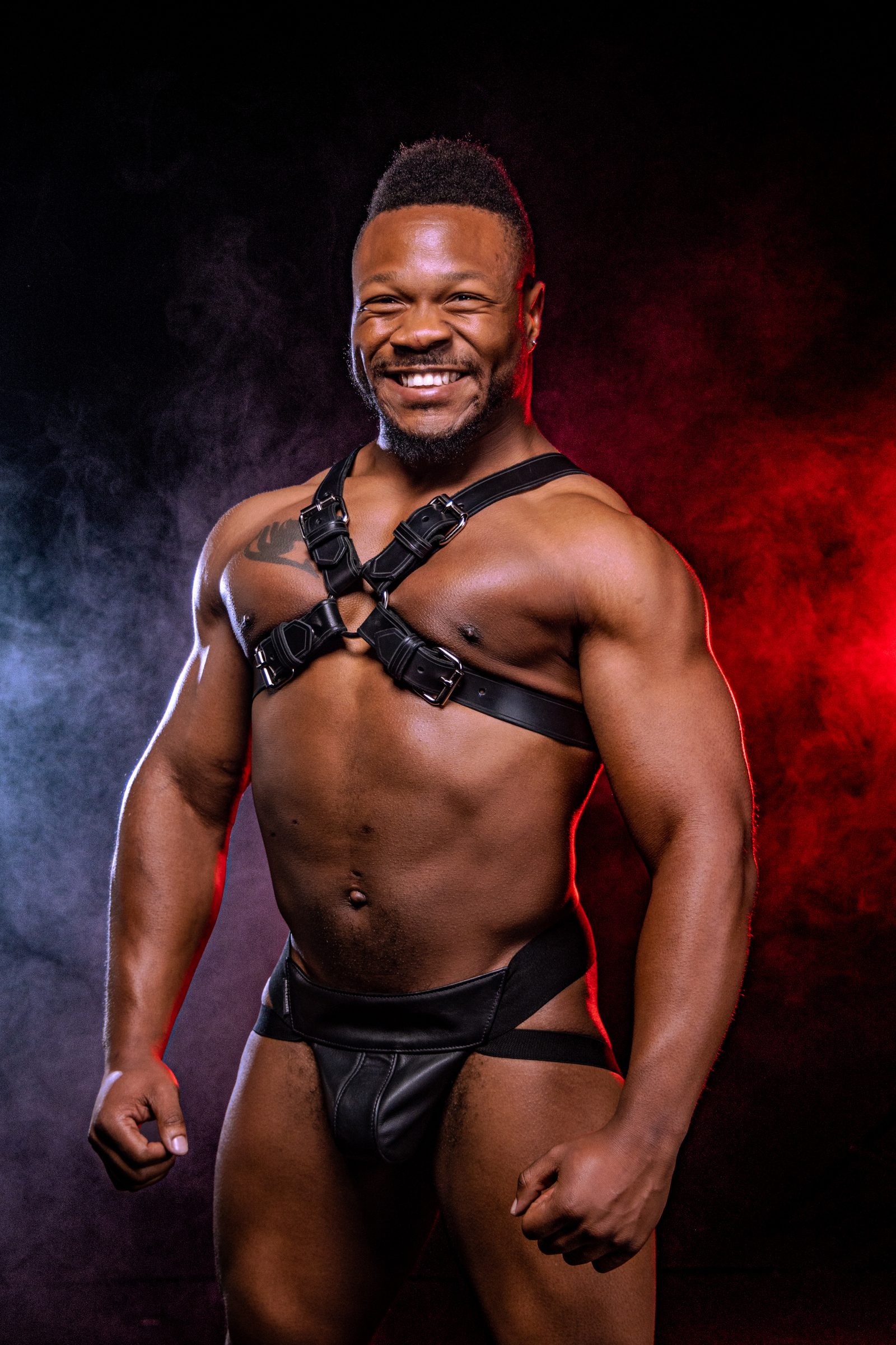 A Black man in a leather harness and jock breaks into a wide, open smile, lit by warm and cool studio smoke that frames him against near-black.