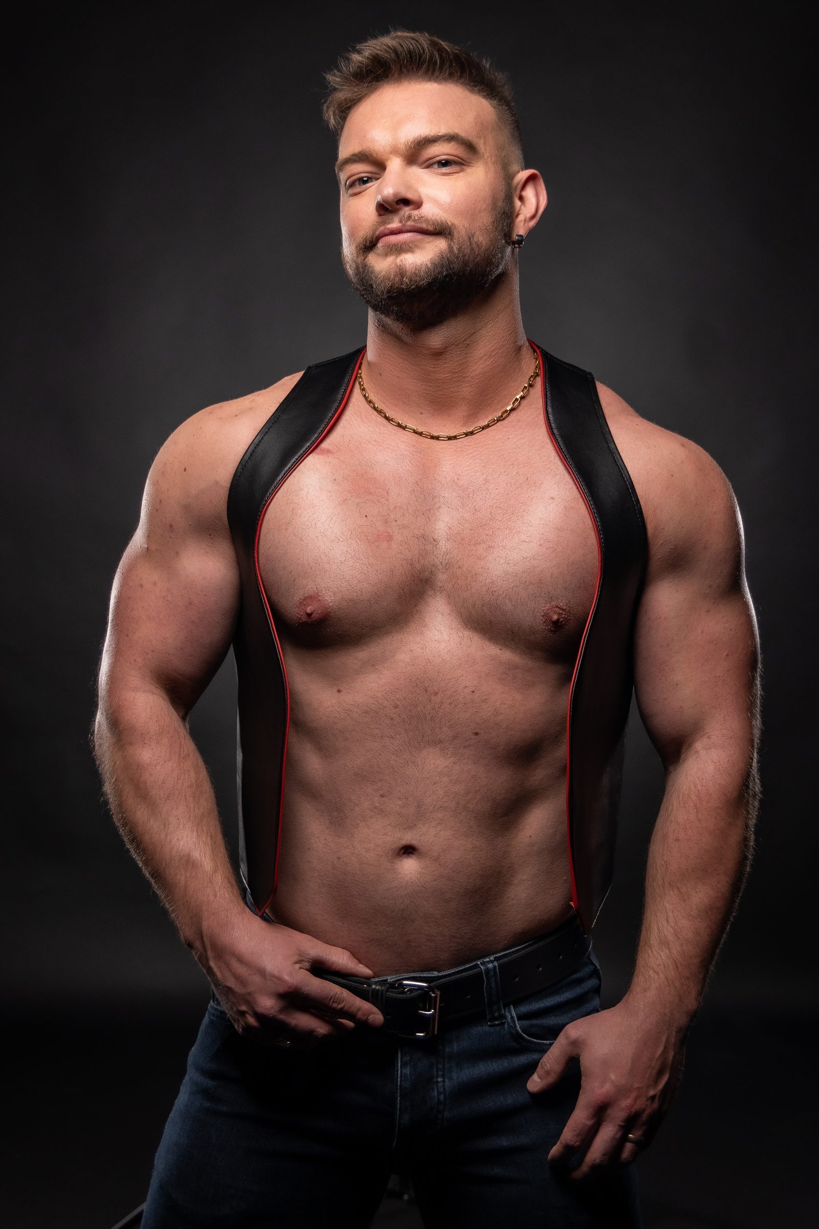 A man in a red-trimmed leather harness vest and jeans rests his hands at his belt against a graphite studio ground, holding a calm, level look.