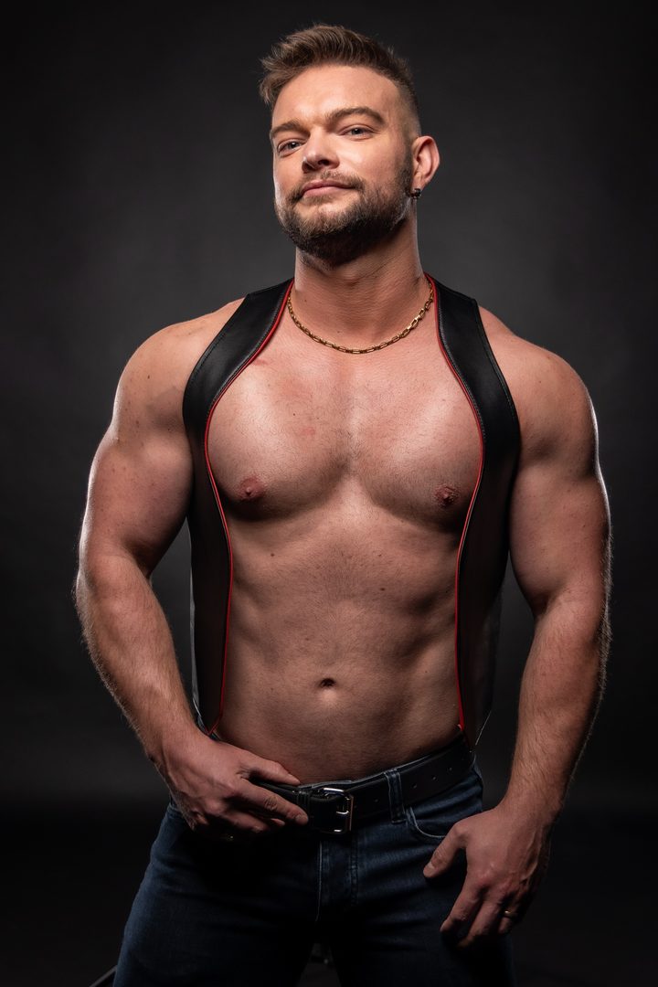 A man in a red-trimmed leather harness vest and jeans rests his hands at his belt against a graphite studio ground, holding a calm, level look.