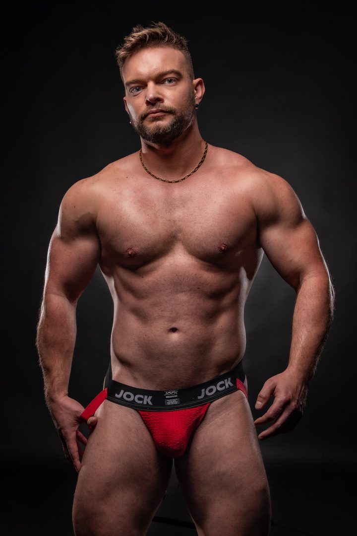 A sandy-haired man in a gold chain and a red jock stands square on a near-black studio ground, meeting the lens with a calm, level look.