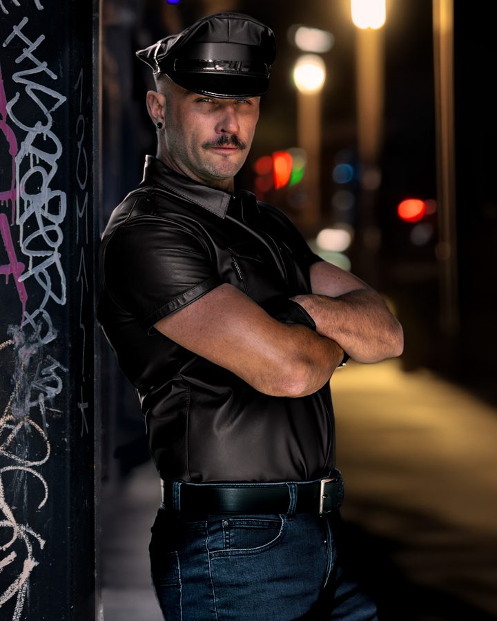 A mustachioed man in a leather cap and short-sleeve leather shirt leans against a graffitied wall with arms crossed, night traffic softened to bokeh behind him.