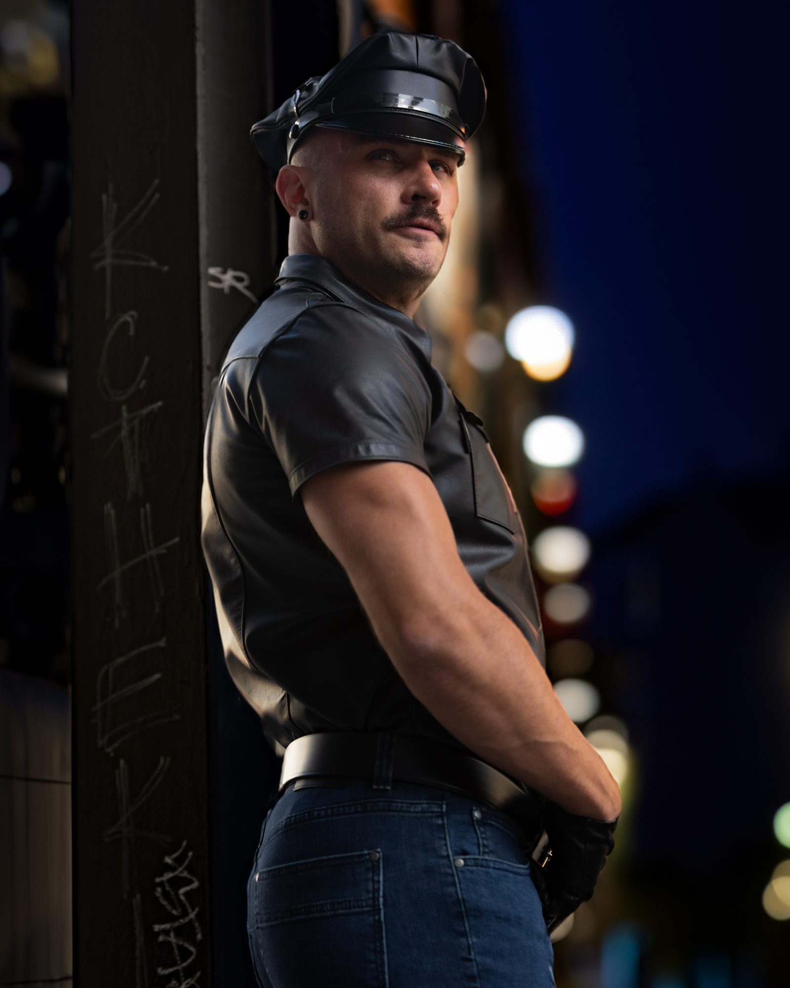 Scene portrait: a man in leather turns three-quarters away on an SF night street, the city lights softened to bokeh behind him.