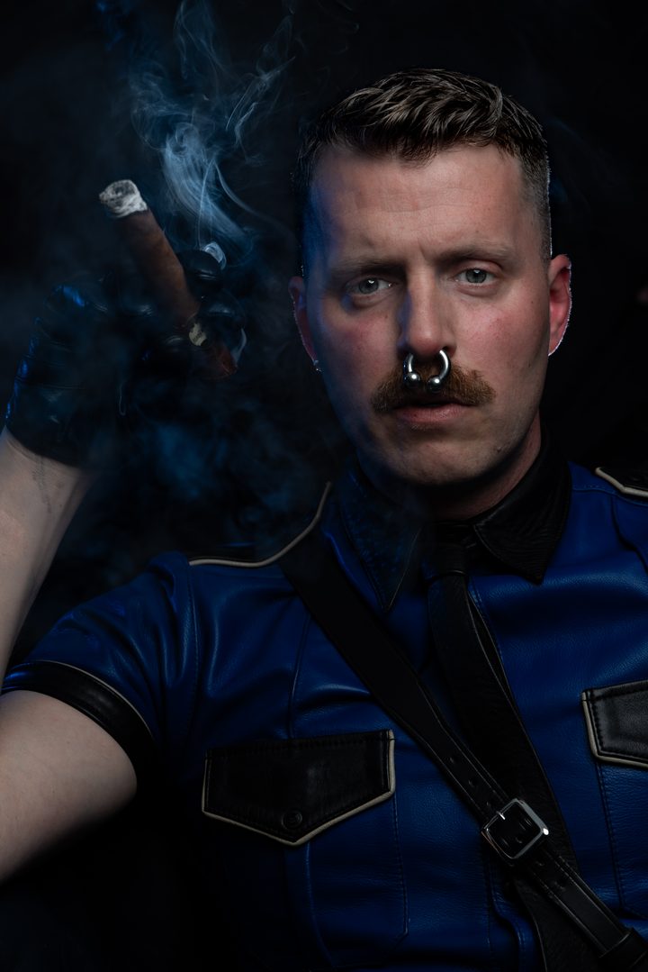 A mustachioed man in a blue leather police shirt raises a cigar through a veil of blue smoke, held against near-black by a single hard strobe.