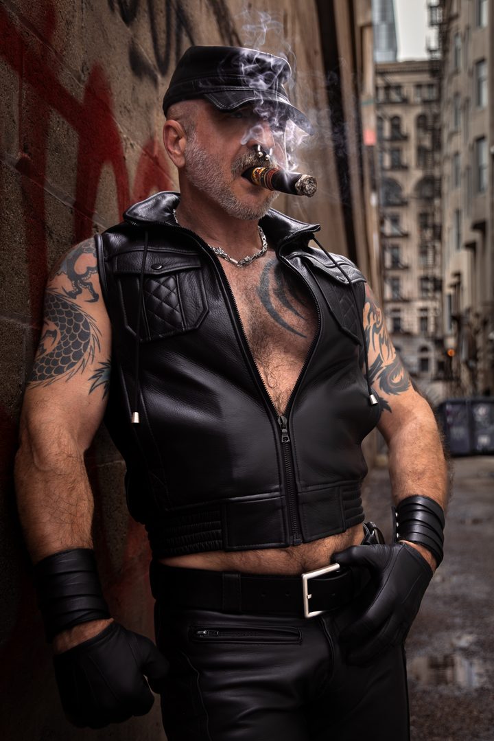 A silver-bearded leather man with a cigar leans against a graffitied San Francisco alley wall, smoke curling past the city behind him.
