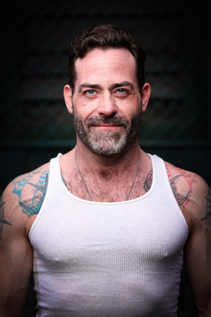 A silver-bearded man in a white tank top, ship and compass tattoos across his shoulders, meets the lens with a warm, settled half-smile.