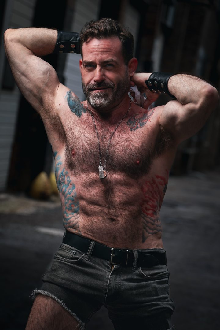 A graying-bearded man in fingerless gloves and dog tags raises both arms behind his head in a gritty alley, the lane falling away soft behind him.