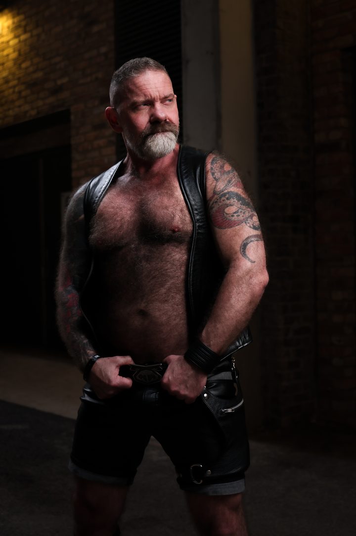 A silver-bearded, broad-bodied man in an open leather vest and shorts stands with hands set at his belt in a dim brick alley, eyes off toward the light.