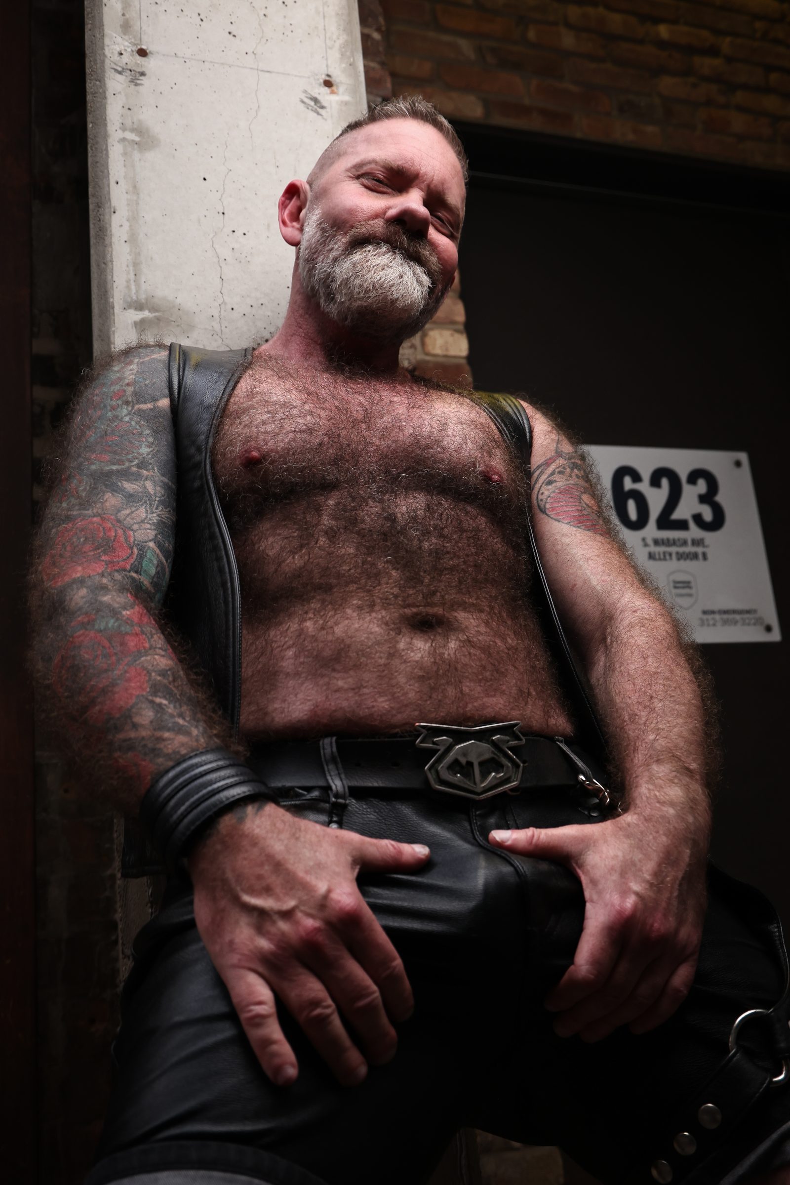 A silver-bearded man in a leather vest and chaps leans into an alley doorway marked 623, tattooed arms framing a calm, self-possessed look.