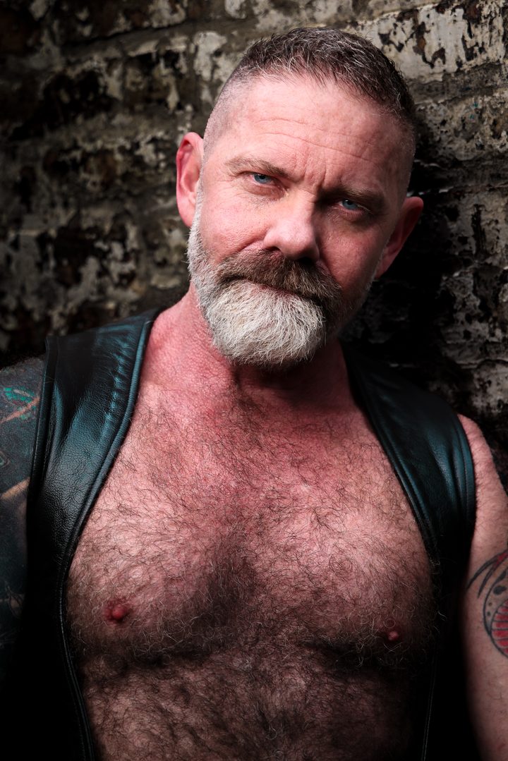 A silver-bearded, broad-chested man in a leather vest holds the lens with an intense gaze against a weathered, paint-flaked stone wall.