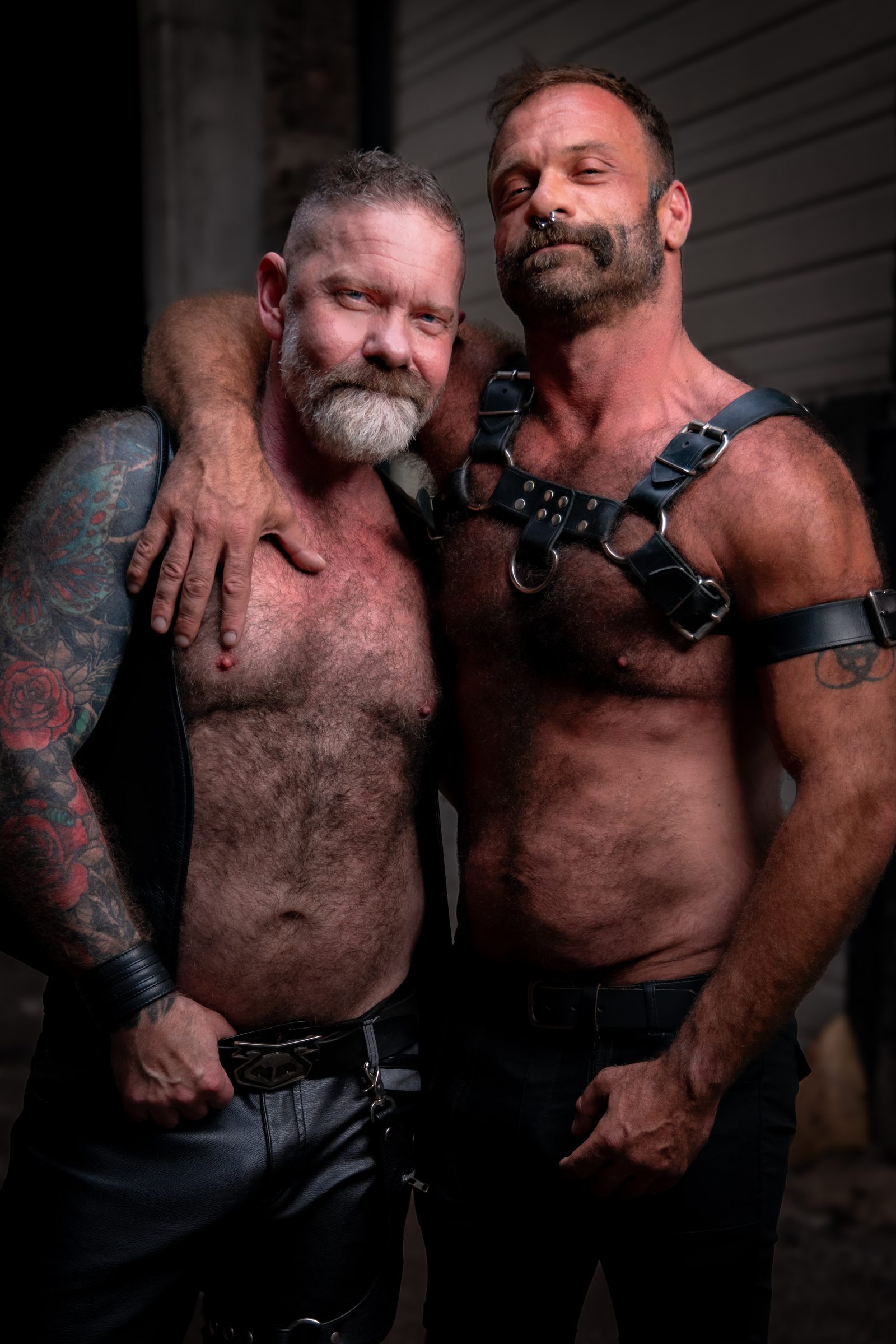 Two leather men, one in a vest and one in a harness, stand close with an arm around each other in a dim warehouse, both meeting the lens.
