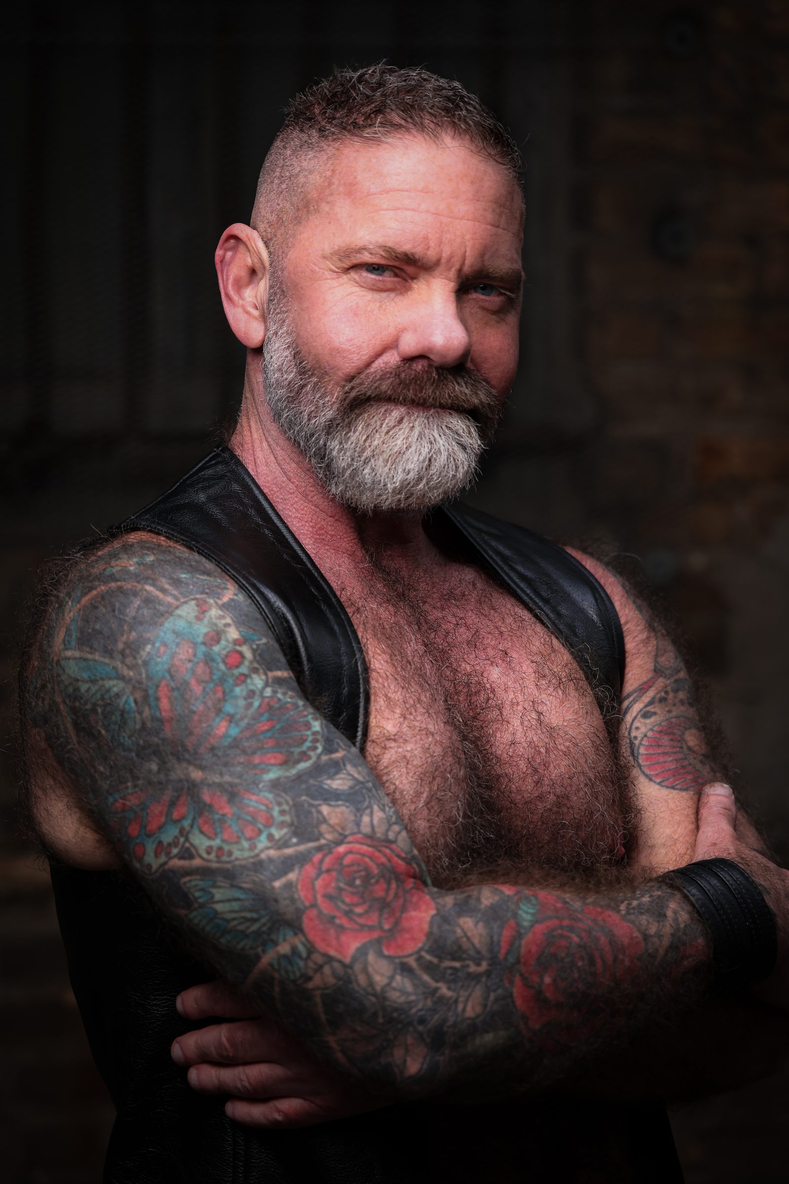 A silver-bearded man in a leather vest folds his rose-and-butterfly tattooed arms and meets the lens with a settled, knowing half-smile.
