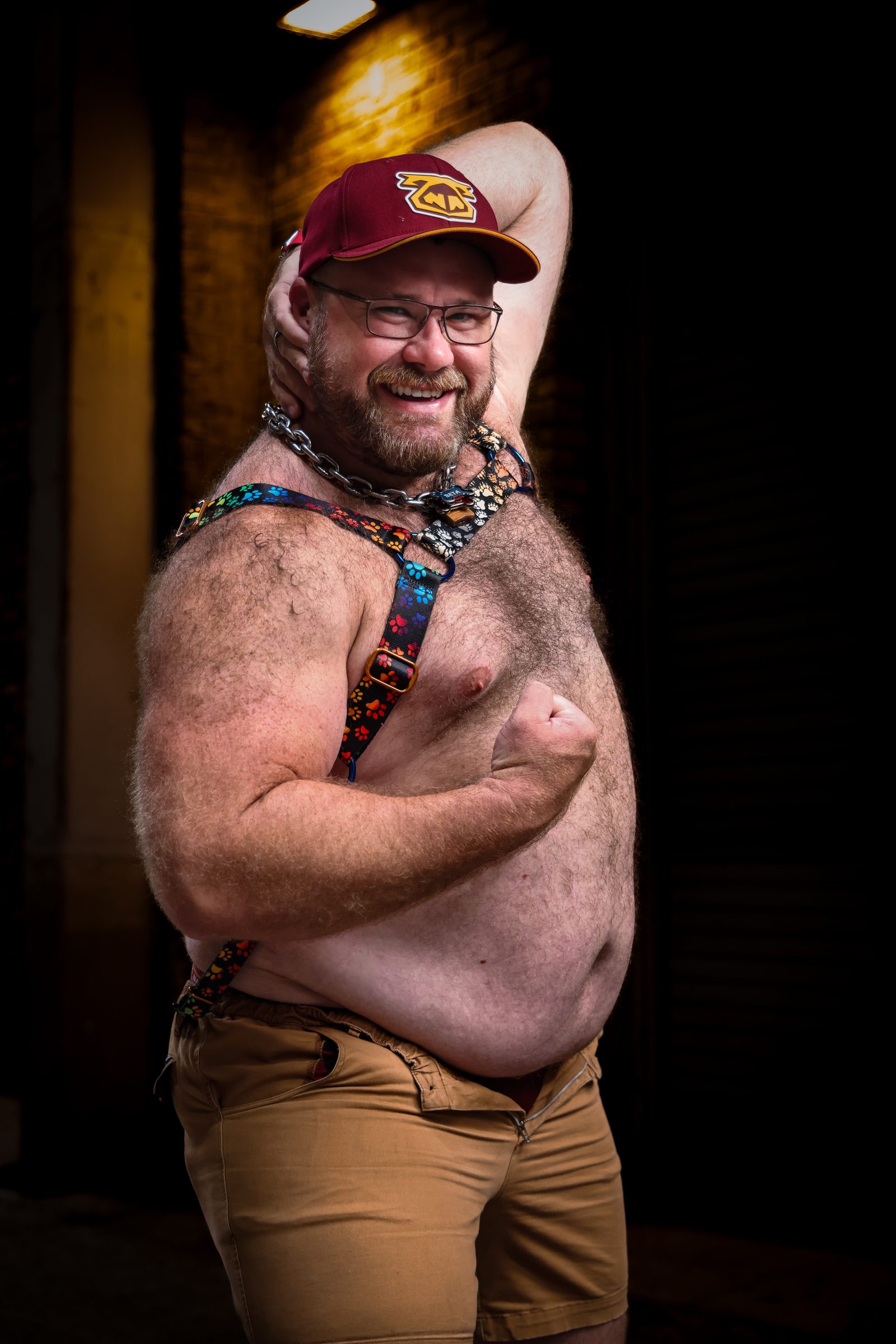 A broad, heavyset bear in a ballcap, glasses, and a floral harness flexes one arm with a delighted grin in a warm brick alley.