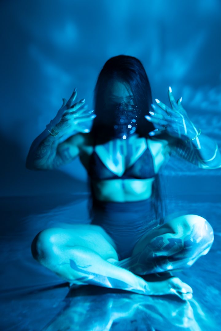 A seated figure lit deep red, a mesh garment and patterned light wrapping the body against a warm, textured ground.