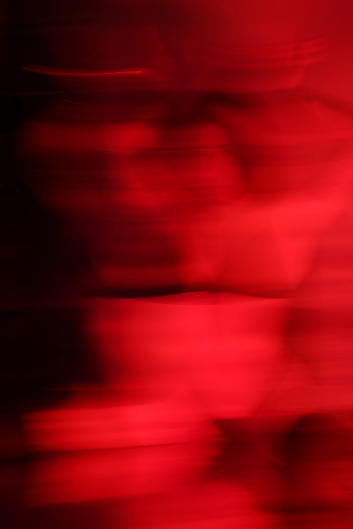 A standing figure pulled into a vertical blur of deep red by a long exposure, the form reduced to motion and color.