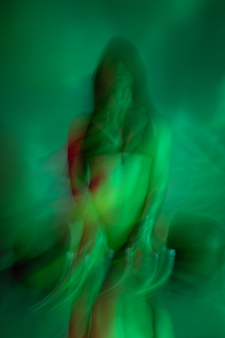 A figure in green and red projected light, the colors layered across the body until the frame reads as overlapping exposures.