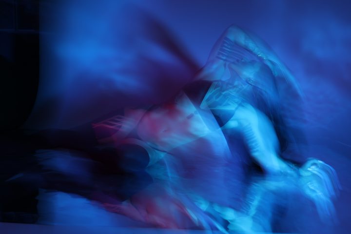 A cobalt-lit figure low in the frame, club light doubling the form in soft blue streaks behind the body.