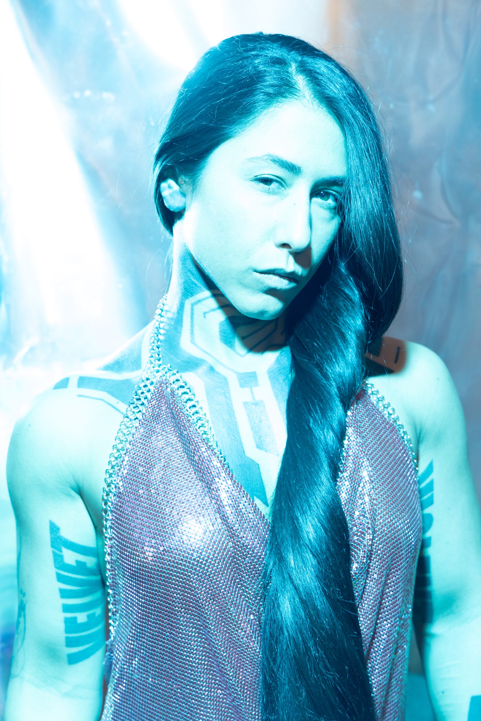 An icy-blue beauty portrait from the Light & Body series, mesh and a long braid, geometric blackwork lit by gelled strobe and held tack-sharp.