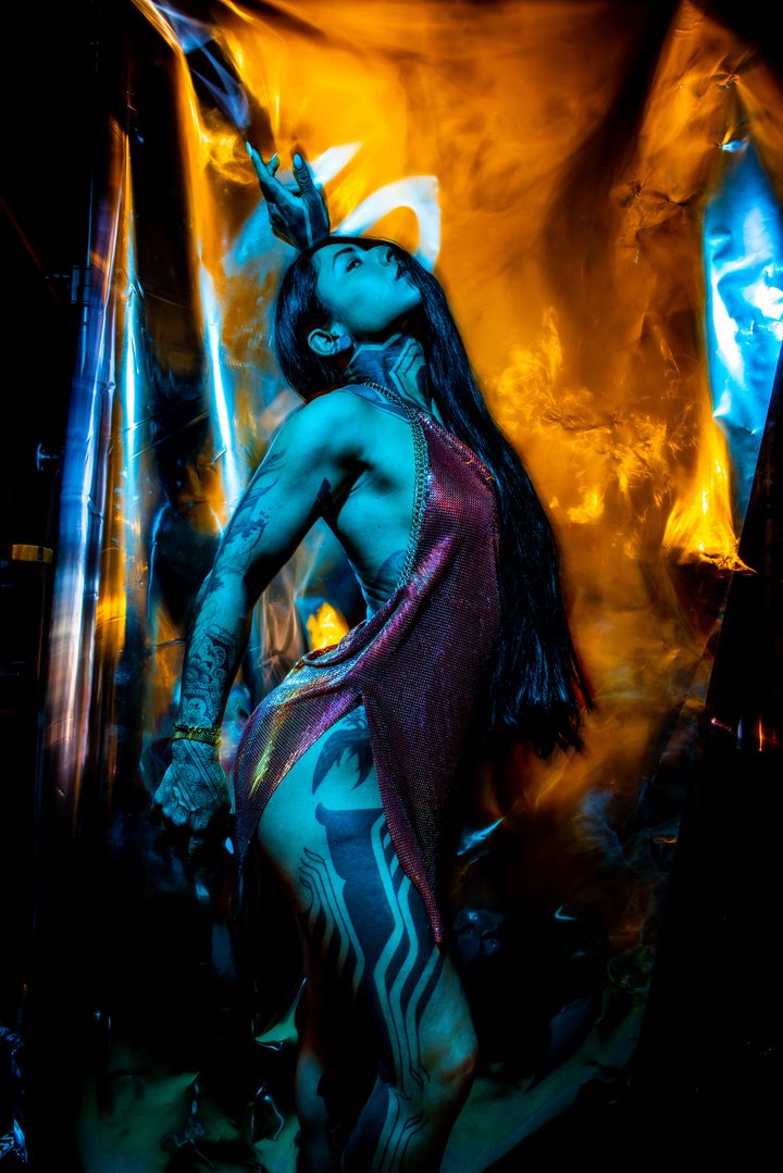 A figure against a warm amber-and-blue ground, draped reflective material catching the gelled light like fire.