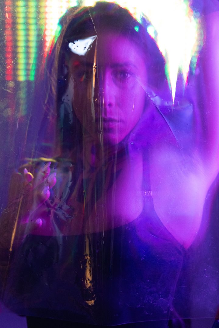 Projected florals mapped over a figure behind scratched acetate, magenta and violet light fracturing the surface.