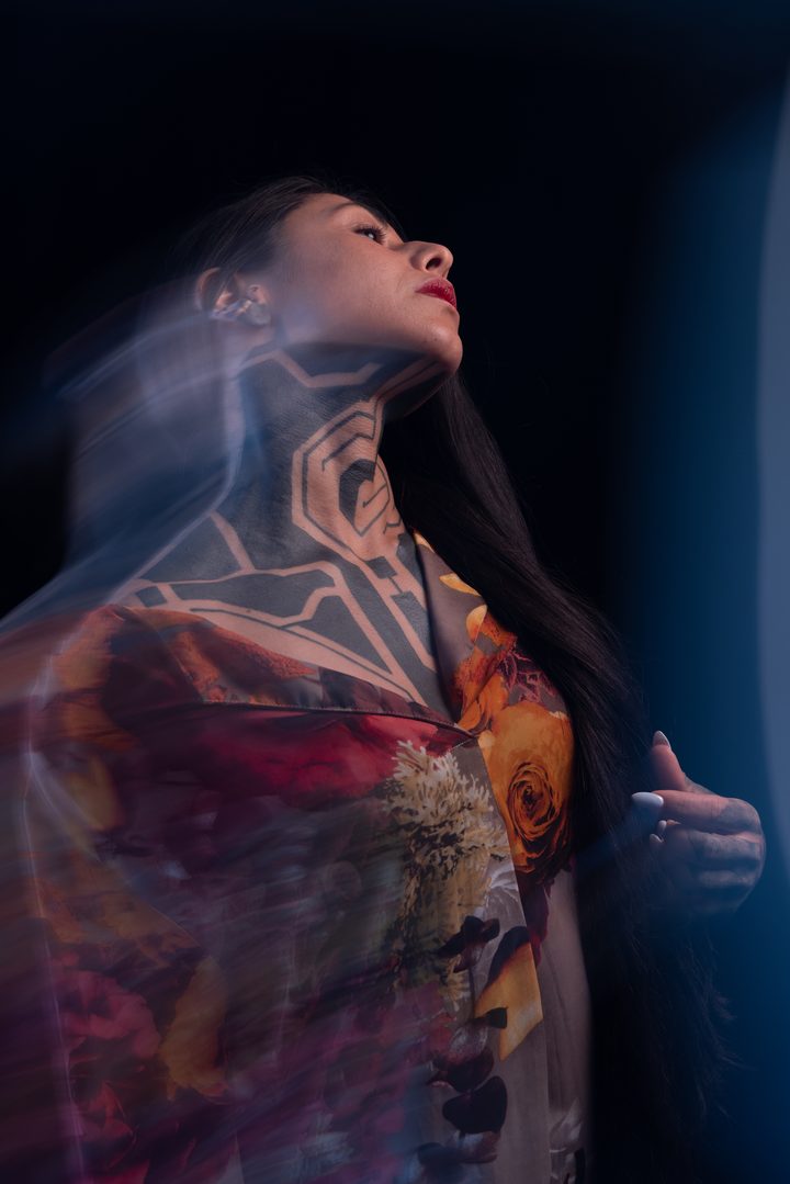 A figure tilting back in a floral garment, projected geometric linework on the neck, trailed by long-exposure motion.